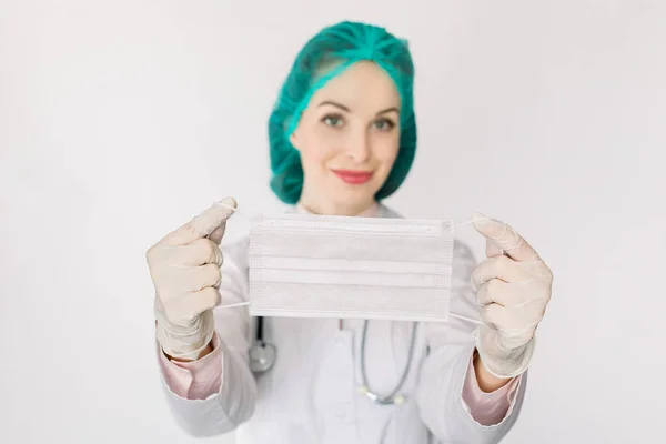 Молода кавказька усміхнена жінка-лікар в шапці, одягнена в захисну маску обличчя від інфекцій, бактерій і вірусів, ізольованих на білому. Зосередься на масці в руках. Coronavirus covid-19 концепція. — стокове фото