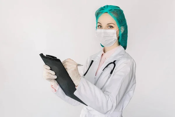 Портрет молодої красивої жінки-лікарки в білій шубці, медичній шапці та масці для обличчя, тримає буфер обміну та запису нотаток, дивлячись на камеру, стоїть на ізольованому білому тлі — стокове фото