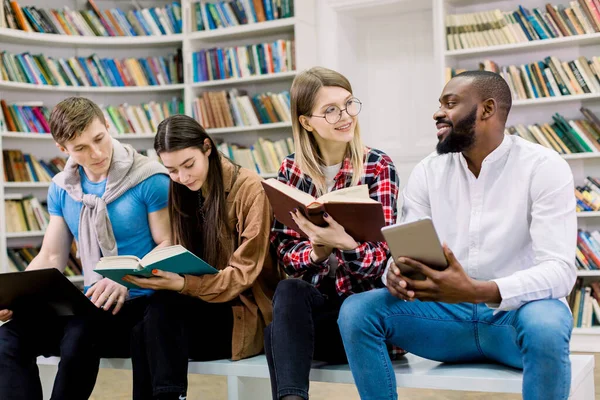 La elección entre libros de papel y educación con aparatos electrónicos. Cuatro estudiantes amigos multirraciales, sentados juntos en la biblioteca universitaria y estudiando, usando libros impresos, tabletas y computadoras portátiles — Foto de Stock