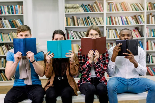 Τέσσερις πολυεθνικοί φίλοι φοιτητές, δύο αγόρια και δύο κορίτσια με καθημερινά ρούχα, να κρύβουν πρόσωπα πίσω από ανοιχτά βιβλία και να κοιτάζουν την κάμερα. Ομάδα φοιτητών με βιβλία στη σύγχρονη πανεπιστημιακή βιβλιοθήκη — Φωτογραφία Αρχείου