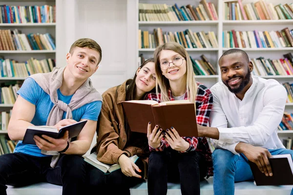 Quatro estudantes multiétnicos alegres amigos na biblioteca, lendo livros, se comunicando e passar o tempo juntos. Feliz Dia Internacional do Livro — Fotografia de Stock