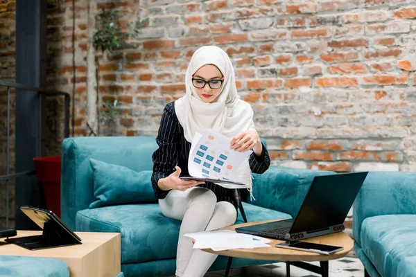 Чудова успішна арабська бізнес-жінка сидить на блакитному дивані перед цегляною стіною і працює з інформацією про проекти на папері та ноутбуці. Мусульманка працює в сучасному офісі. — стокове фото