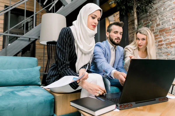 Троє молодих багатоетнічних колег, кавказький чоловік і жінка, мусульманка в Хіджабі, які розмовляють і користуються ноутбуком, сидять на блакитному дивані в сучасному офісі. — стокове фото