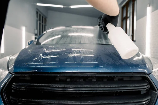 Menutup tangan pekerja cuci mobil laki-laki dalam sarung tangan karet hitam, membersihkan kap mobil biru dengan botol semprotan, menempatkan bahan lilin dingin di permukaan. Pemeliharaan mobil dan rincian konsep — Stok Foto