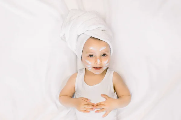 Bambina bambina che si diverte dopo il bagno con asciugamano sulla testa e crema viso su guance, naso e fronte, sdraiata su sfondo bianco. Igiene del bambino, cura del viso, spa e bagno — Foto Stock