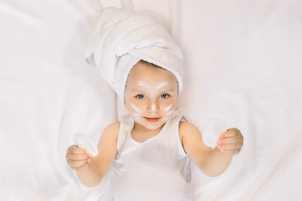 Bella bambina di 2 anni che si rilassa nella camera da letto dopo la doccia, sdraiata sul letto, indossa un asciugamano bianco sulla testa e tiene in mano due batuffoli di cotone. Cura del mattino e igiene del bambino — Foto Stock