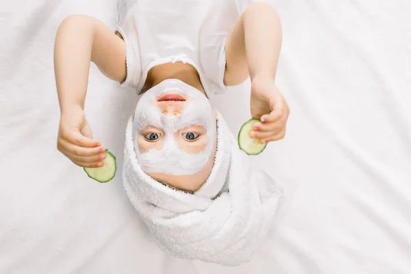 Zabawna dziewczynka w masce na twarz i ręcznik na głowie, trzymająca dwa kawałki świeżego ogórka i patrząca w kamerę, leżąca na białym tle po kąpieli — Zdjęcie stockowe