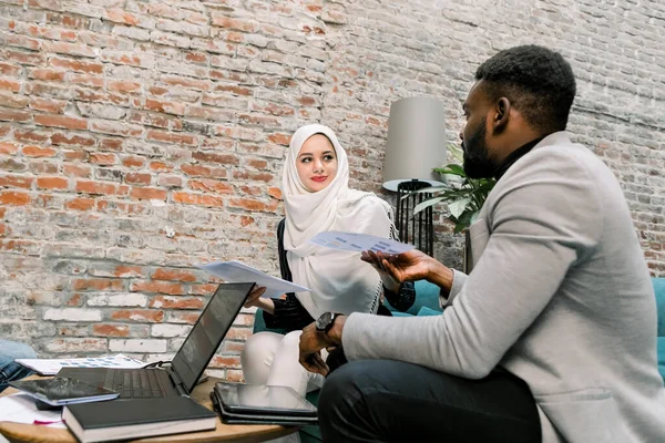 Зображення багатонаціональних ділових партнерів, гарний бородатий африканський чоловік і досить мусульманська жінка в Хіджабі, сидячи в офісній кімнаті біля цегляної стіни, працюючи з паперами і обговорюючи ідеї — стокове фото