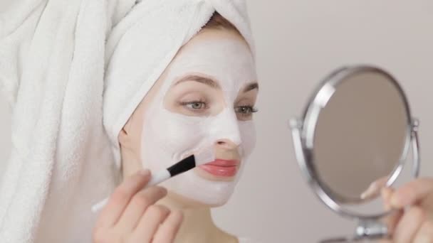 Sorrindo jovem encantadora mulher com toalha na cabeça aplicando máscara de barro cosmético branco no rosto usando maquiagem escova e olhando para o espelho. Close up tiro, isolado no fundo branco — Vídeo de Stock