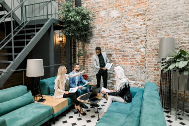 Genç, koyu tenli, finans projesinin sonuçlarını gösteren genç bir işadamı, elinde çok ırklı iş arkadaşlarına ait grafiklerle rahat koltuklarda oturuyor.