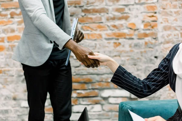 Business concept of hand shaking. Cropped image of shaking hands after agreement between business lady in black jacket and dark-skinned businessman in the modern loft office room