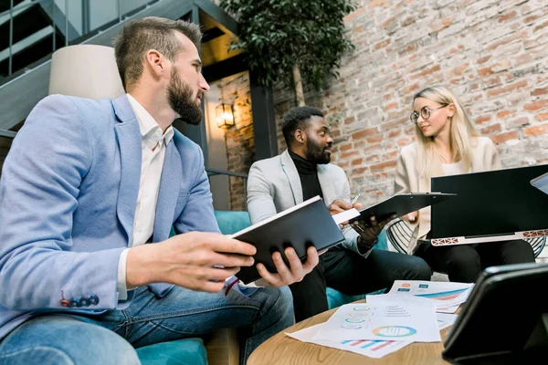 Große Entscheidungen zu treffen. Junge kaukasische blonde Frau und zwei Männer, kaukasische und afrikanische, gestikulierend und lächelnd am Bürotisch sitzend vor Backsteinmauer — Stockfoto