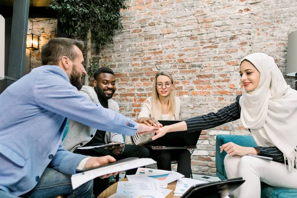Успішна багатоетнічна команда розумних бізнесменів святкує успіх нового проекту і складає руки в центрі над столом в сучасному лофт офісі — стокове фото