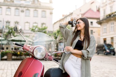 Moda gözlüklü, sıradan giyinen, elinde retro kamera olan ve eski Avrupa şehir binalarının arka planında kırmızı motorsikletin üzerinde oturan genç ve güzel bir kız. Kentsel yaşam tarzı