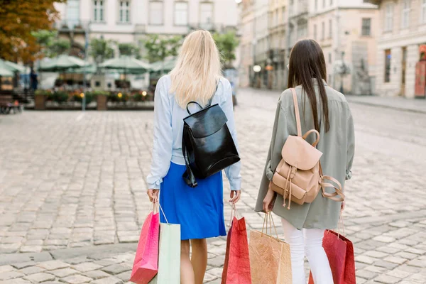 Vista trasera de dos guapas mujeres rubias y morenas jóvenes con mochilas de cuero y coloridas bolsas de compras caminando juntas en la plaza en el viejo centro de la ciudad europea. Feliz fin de semana de compras — Foto de Stock