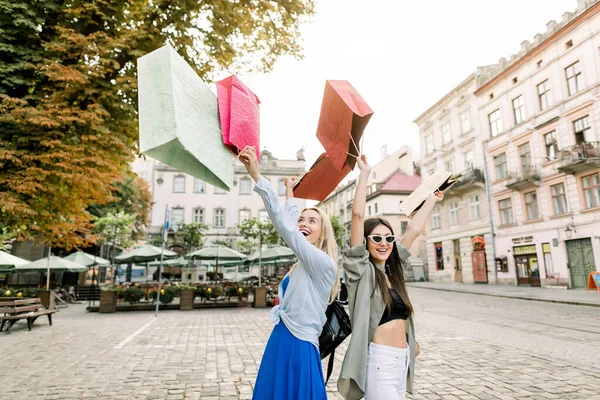 Two pretty young Caucasian girls, blond and brunette, wearing casual clothes, strolling on the street with arms raised, holding shop packages and enjoying their walk in beautiful European city center — Stock Photo, Image