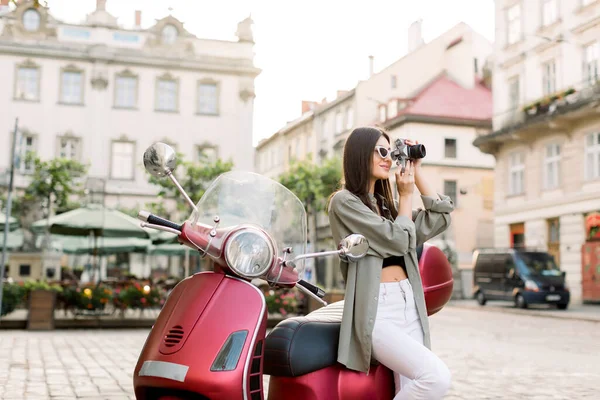 Aantrekkelijke brunette jonge vrouw in stijlvolle kleding en zonnebril maken van foto 's van prachtige architectuur gebouwen, leunend op de rode scooter op de achtergrond van de oude stad straat — Stockfoto