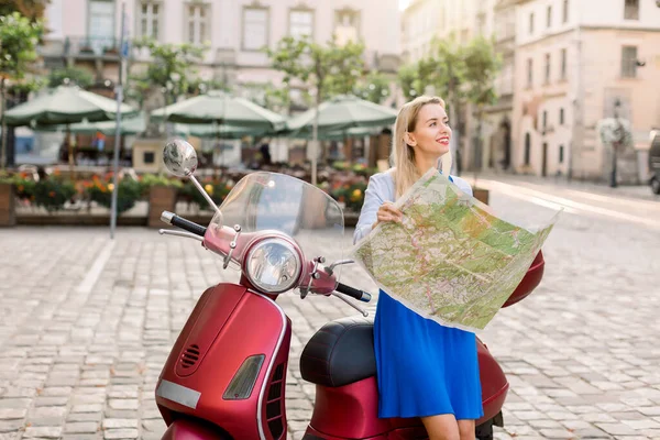 Mooie blonde vrouw in blauwe jurk zit op rode scooter en houdt toeristische kaart, terwijl reizen en genieten van haar zomervakantie. Reisconcept met kaart in de Europese stad — Stockfoto