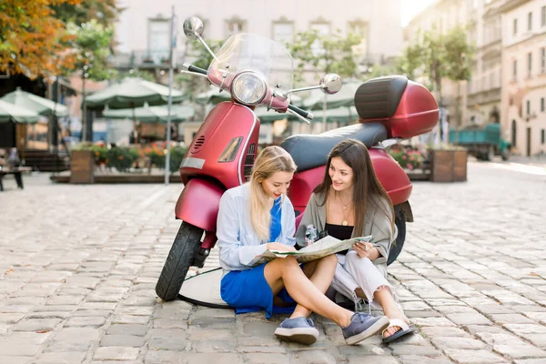 Urban style portrait of two joyful young girls sitting on the pavement near modern red motor bike scooter and using touristic city map, looking for interesting places to visit. City tourism concept — Stock Photo, Image