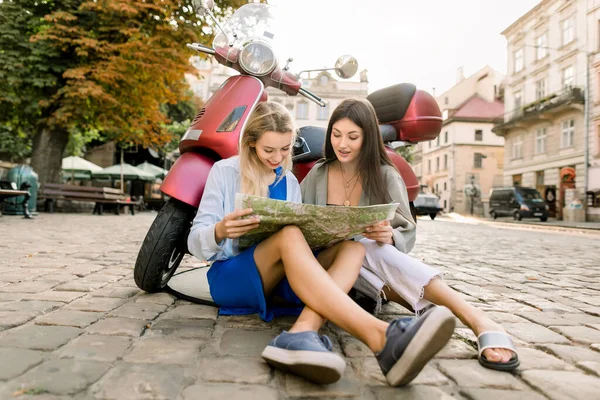 Two friends pretty Caucasian women sitting on the pavement leaning on red scooter and holding city map to find famous places and buildings, looking for a road. Vacations in old European city — Stock Photo, Image