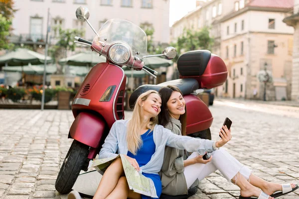 Two charming smiling young women in casual clothes sitting on the street pavement outdoors near stylish red motor biker and having fun making selfie photo on smartphone on the background of old city — Stock Photo, Image