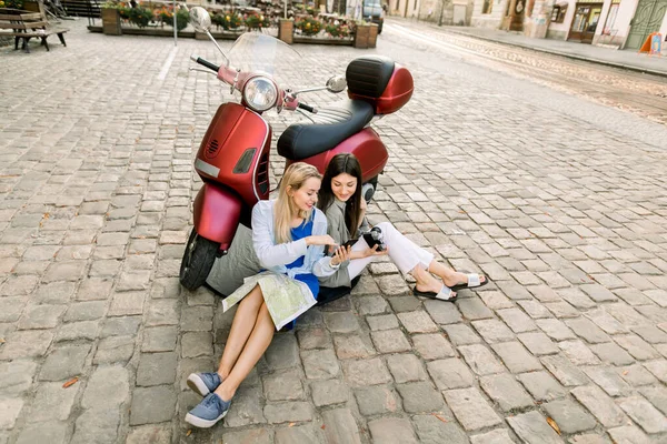 Портрет двух кавказских подруг в городе, сидящих на тротуаре с туристической картой, фотоаппаратом и опирающихся на красный скутер, в поисках интересных мест — стоковое фото