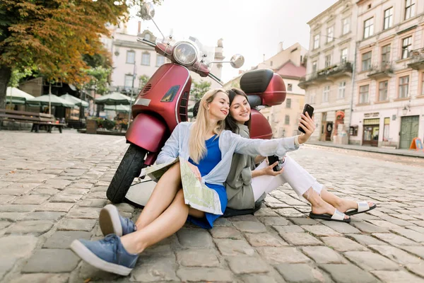 Two charming smiling young women in casual clothes sitting on the street pavement outdoors near stylish red motor biker and having fun making selfie photo on smartphone on the background of old city — Stock Photo, Image