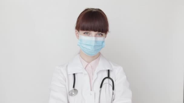幸せな笑顔の女性医師は、科学者の白いコートを着て、聴診器や医療面マスクの白い背景にカメラを見て、ウインクや親指をジェスチャーを表示します。covid-19パンデミックを阻止する — ストック動画