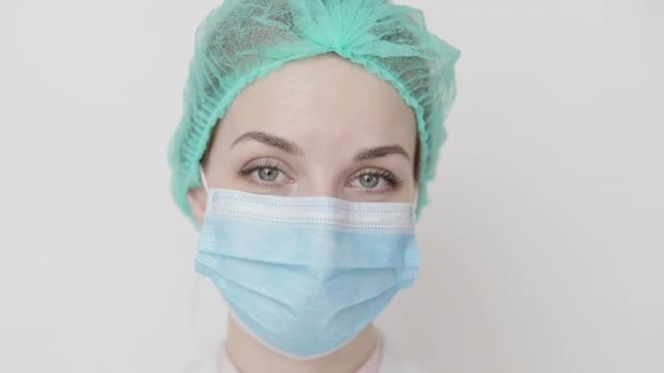 Primer plano de una cara de cirujana, con gorra y máscara médica, guiñando un ojo a la cámara. Enfermera médica profesional que usa gorra de exfoliante, máscara facial protectora, guiño a la cámara — Vídeos de Stock