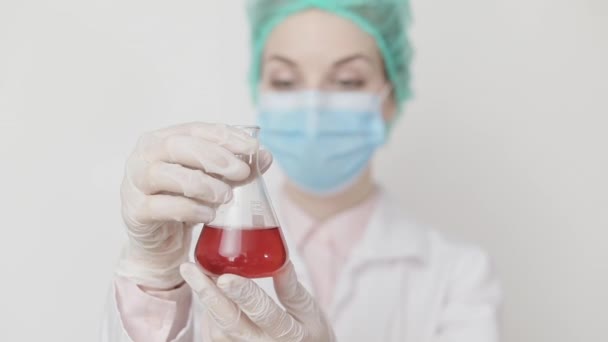Retrato de una doctora científica o asistente de laboratorio vestida con bata blanca, máscara y gorra, sosteniendo el frasco con líquido rojo, mirándolo y agitándolo. Aislado sobre blanco. Concéntrate en el frasco — Vídeos de Stock