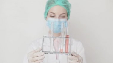 Genç kadın doktor, bilim adamı, eczacı, laboratuvar teknisyeni ellerinde koronavirüs için kan testi olan test tüpleri tutuyor. Koruyucu maske, kep ve lateks eldiven takan bir kadın test tüpleri tutuyor.