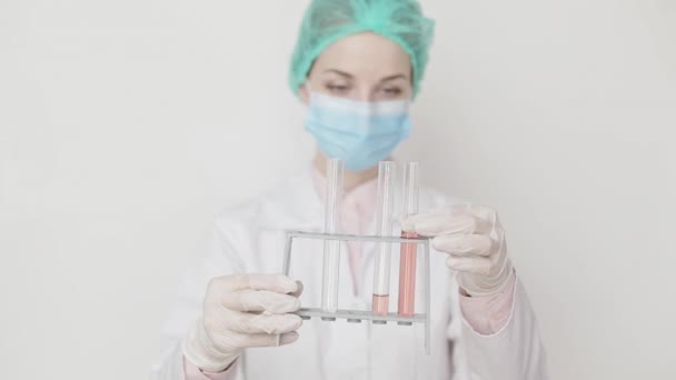 Młoda atrakcyjna kobieta naukowiec lub lekarz w białej powłoce, czapce, masce i rękawiczkach trzymających szklane probówki z próbkami testowymi i patrząc na jedną z probówek, stojących na białym tle. — Wideo stockowe