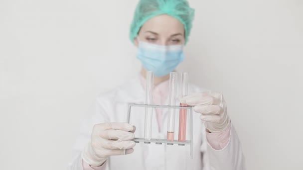 Joven científica atractiva o doctora en bata blanca, gorra, máscara y guantes sosteniendo tubos de ensayo de vidrio con muestras de prueba y mirando uno de los tubos, de pie sobre fondo blanco . — Vídeos de Stock