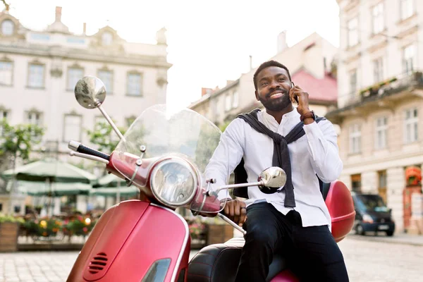 Knappe bebaarde Afrikaanse zakenman geniet van zijn succesvolle telefoongesprek, terwijl hij op de moderne rode motor zit op de achtergrond van oude stadsgebouwen. Bedrijfsconcept — Stockfoto