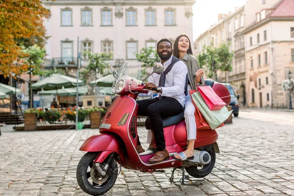 Jonge mooie multiraciale paar, Afrikaanse man en blank meisje, rijden op rode motor op de straat van de stad, gelukkige vrouw houdt boodschappentassen. Zomer Europa vakantie, reizen concept — Stockfoto