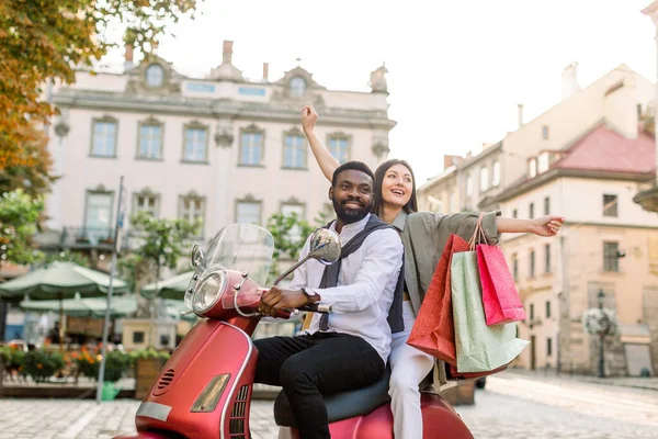 Веселая гламурная молодая многонациональная пара на красном винтажном скутере на улице, африканский мужчина носит белую рубашку и брюки, а белая женщина держит красочные сумки с дорожными сувенирами — стоковое фото