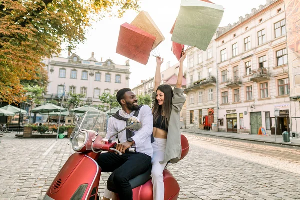 Charmante glimlachende blanke vrouw zit achter haar knappe Afrikaanse vriendje op de rode scooter met winkeltassen in haar handen opgeheven op de achtergrond van Europese stadsgebouwen — Stockfoto