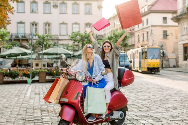 Pretty fashionable smiling young Caucasian women in stylish sunglasses sitting on the red motor bike scooter, holding colorful shopping bags and having fun on street square. Shopping and travel — Stock Photo, Image