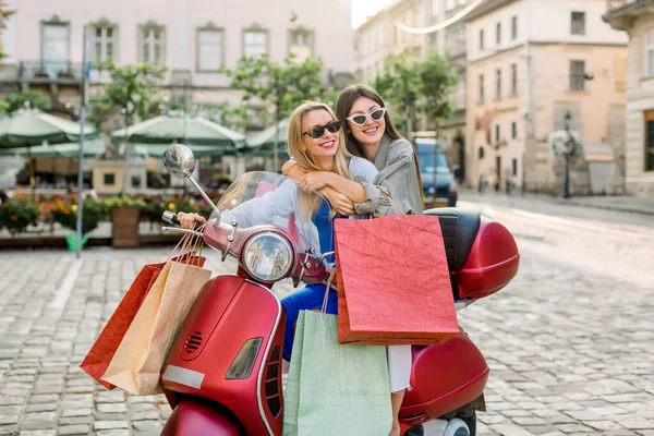 Two attractive young girls best friends wearing casual clothes and sunglasses sitting on the red scooter with a lot of shop bags, on the background of the ancient buildings in old European city — Stock Photo, Image