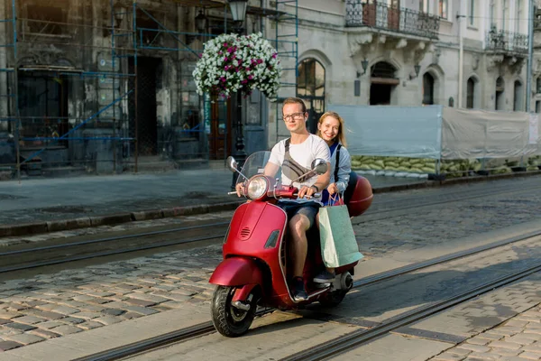 Стильные белый мужчина и женщина с мотоциклом. Счастливая пара едет на красный мотор в городе, радостная блондинка в синем платье, держа много красочных мешков для покупок. Концепция путешествия и покупок — стоковое фото
