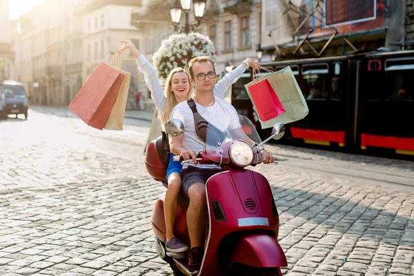 Gelukkig lachend mooi meisje met winkeltassen in haar handen zittend achter knappe man op de rode scooter en rijdend door de straat van de stad. Stedelijke levensstijl en winkelconcept — Stockfoto