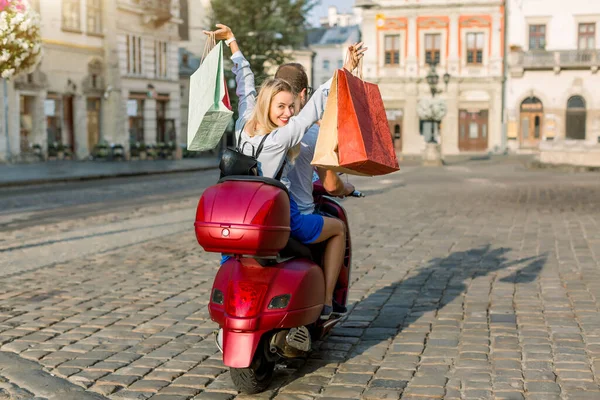 Back view of young couple riding red scooter on a sunny day in old European city. Pretty happy blond woman with shopping bags smiles and looks at camera. Travelling with motor bike, urban tourism — Stock Photo, Image