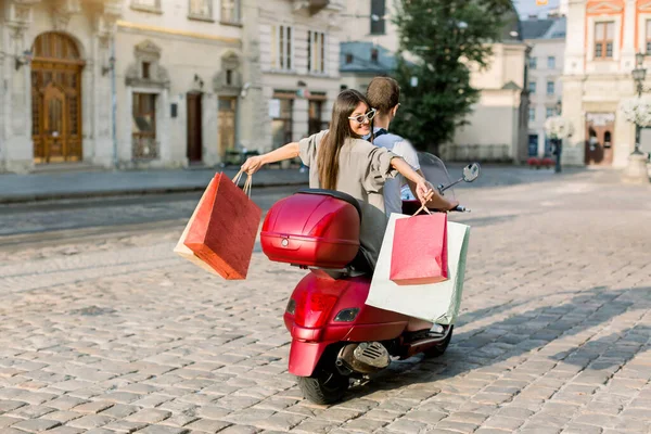 Bakc view of pretty brunette young lady in stylish sunglasses with many shopping bags in hands riding on red scooter with her boyfriend through the city street. Travelling and shopping concept — Stock Photo, Image