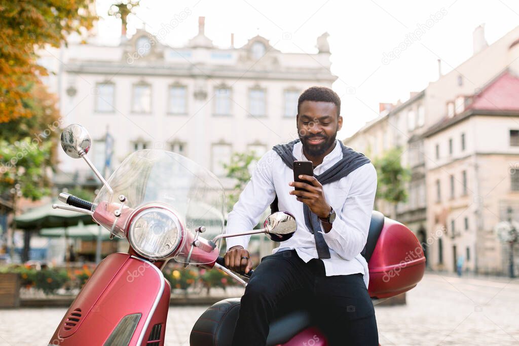 Portrait of smiling young bearded african american guy in smart casual wear using his smartphone, while sitting on the red scooter outdoors on the old city square. Urban lifestyle, business