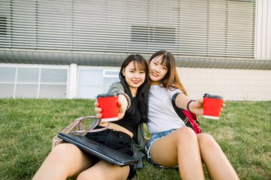 Güzel Asyalı bayan üniversite öğrencileri ellerinde kahve fincanları tutuyorlar ve onları kameraya gösteriyorlar, modern şehir binasının önündeki yeşil çimlerin üzerinde oturuyorlar. İnsanlar, dostluk ve rahatlık