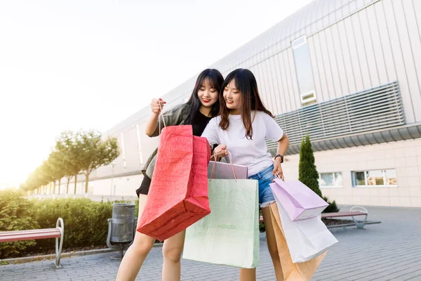 Two happy smiling charming 25s asian women showing each other their shopping bags inside, standing on the shopping store building background outdoors in the city. Travel and shopping concept — Stock Photo, Image