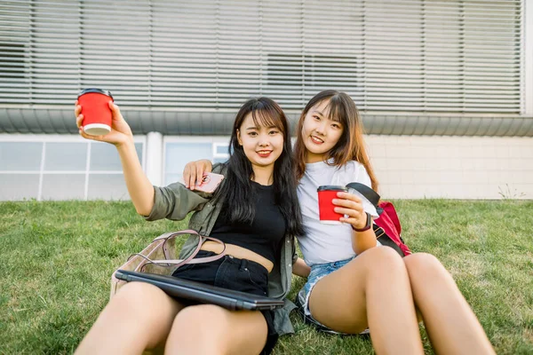 Dos guapas chicas asiáticas morenas disfrutando de su tiempo juntas, sentadas al aire libre sobre hierba de pasto en la ciudad, posando ante la cámara con tazas rojas de café. Ocio, estilo urbano y concepto de amistad —  Fotos de Stock