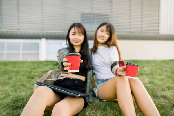 Hermosas estudiantes universitarias asiáticas sosteniendo tazas de papel rojo con café y mostrándolas a la cámara, sentadas al aire libre sobre hierba verde frente a un edificio urbano moderno. Concéntrate en las copas —  Fotos de Stock