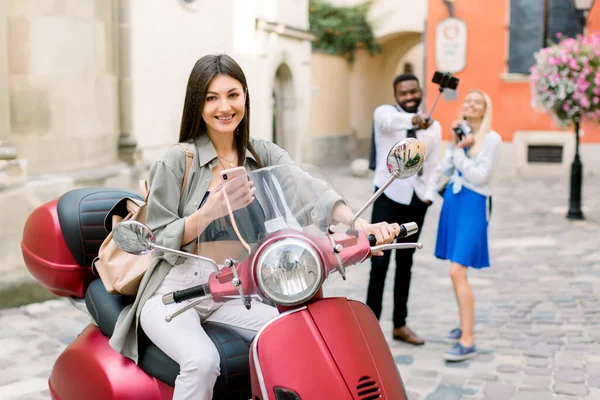 Bella allegra ragazza caucasica bruna seduta sul motorino rosso nella vecchia strada della città, mentre i suoi amici multirazziali, ragazzo africano e ragazza caucasica si divertono e fanno foto di selfie — Foto Stock