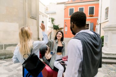 Üç mutlu, gülümseyen çok kültürlü insan, esmer tenli adam ve iki beyaz kadın, eski şehir caddesinde buluşurken beşlik çakıyorlar. Esmer kız kırmızı scooter 'da oturuyor.